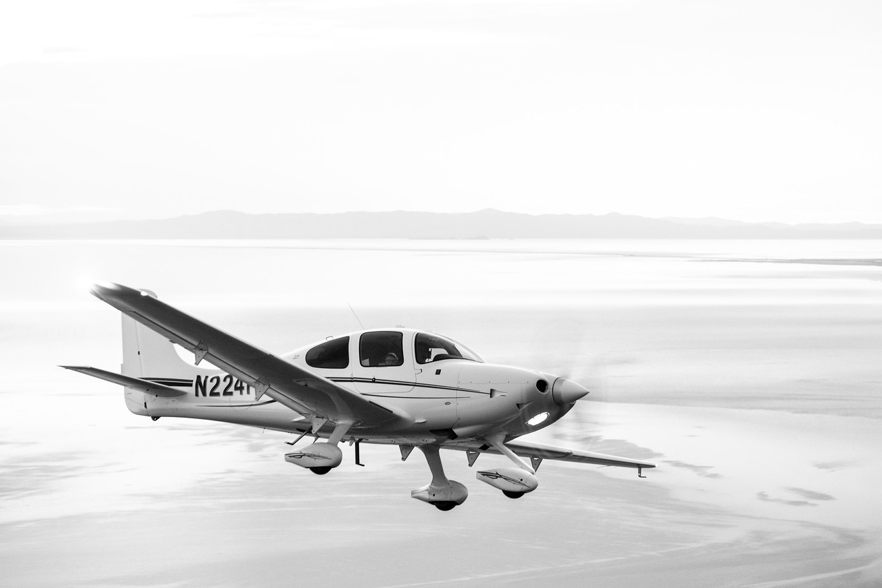 Black-and-white photograph of a Cirrus flying above a pale shoreline.